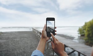 Hand håller upp en svart mobil med kamera läget på och ska ta en bild av stranden som synns bakom, med en lång brygga. 