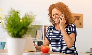 Kvinna som sitter framför en vit laptop och pratar i mobiltelefon. I förgrunden syns en krukväxt och i bakgrunden  en bokhylla.