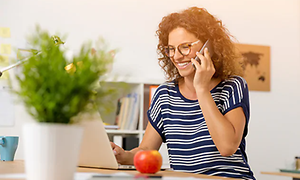 Kvinna som sitter framför en vit laptop och pratar i mobiltelefon. I förgrunden syns en krukväxt och i bakgrunden en bokhylla.