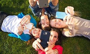Fyra tjejer som ligger ner på en gräsmatta och sträcker upp sina mobiltelefoner mot himlen för att at selfies eller groupies. 