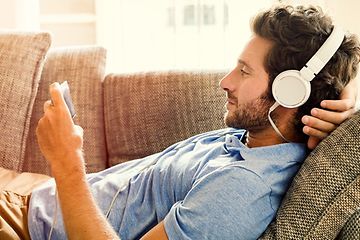 Man ligger i soffan och tittar på något underhållande på sin mobiltelefon, med stora vita hörlurar inkopplade. 