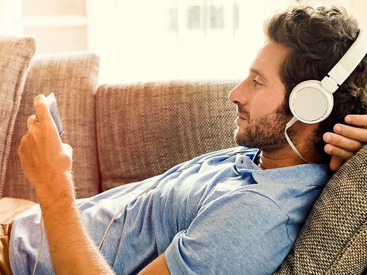 Man ligger i soffan och tittar på något underhållande på sin mobiltelefon, med stora vita hörlurar inkopplade.
