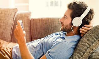Man ligger i soffan och tittar på något underhållande på sin mobiltelefon, med stora vita hörlurar inkopplade. 