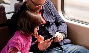 En liten flicka och en äldre man sitter och kollar på något spännande på en mobiltelefon, vid fönstret på ett tåg. 