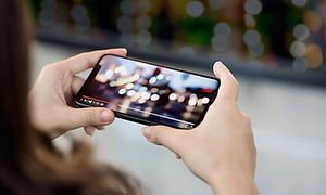 Bild på en mobilskärm som spelar upp en video, man ser baksidan på ett huvud med långt hår och två händer som håller telefonen. 