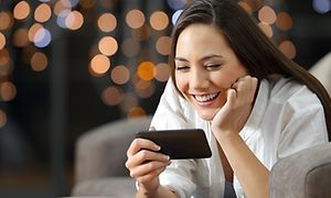 En kvinna som sitter i en fåtölj och är djupt försjunken i det som sker på mobilskärmen, hon ser glad ut.