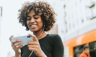 Kvinna som står ute på en gata med en vit mobil i händerna och headset i öronen med fullt fokus på det som sker på skärmen. 
