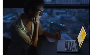 Kvinna i mörkt rum sitter framför en Lonovo Thinkbook med skärm som lyser upp och tangenter med bakgrundsljus. 