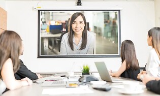 Flera personer sitter på ett möte och en kvinna deltar via video.