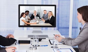 Fyra personer sitter samlade i ett mötesrum och ser mot en skärm där fem andra personer är samlade, till videokonferens. 