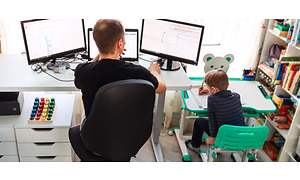 Man som sitter hemma vid ett skrivbord och arbetar, med ett mindre skrivbord bredvid där en pojke sitter och tecknar.