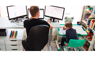 Man som sitter hemma vid ett skrivbord och arbetar, med ett mindre skrivbord bredvid där en pojke sitter och tecknar.