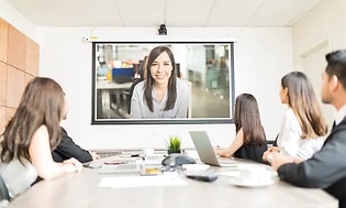 Mötesrum med fem personer som deltar i ett videomöte, en person är ansluten online och syns på en stor bildskärm. 
