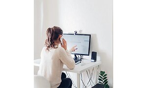 Kvinna sitter vid skrivbord hemma och pratar i telefon, hon pekar på dataskärmen som står på skrivbordet. 