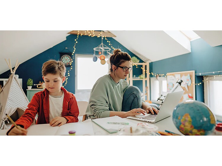 Kvinna sitter på barnrum och arbetar på en laptop med sonen bredvid som sitter och målar. 