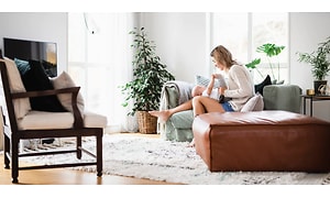 Kvinna sitter i en grön soffa i ett vardagsrum med en liten bebis i knät. Stora ljusa fönster i bakgrunden och andra möbler. 