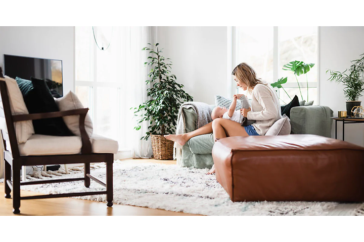 Kvinna sitter i en grön soffa i ett vardagsrum med en liten bebis i knät. Stora ljusa fönster i bakgrunden och andra möbler. 