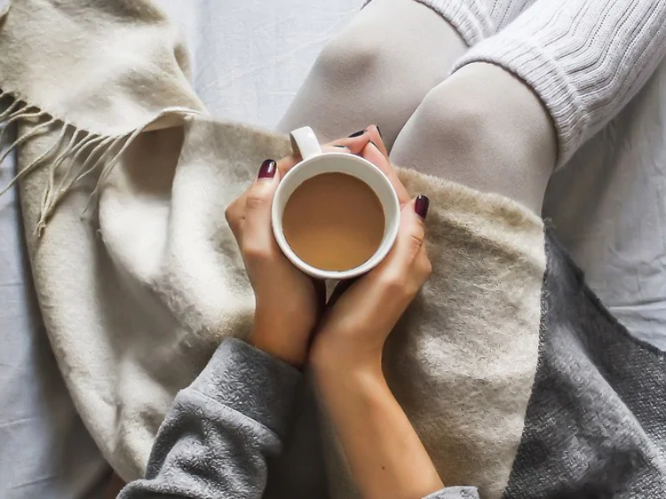 Kvinna sitter med raggsockar och en massa filtar i en säng och har händerna runt en kopp kaffe. Bilden är tagen ovanifrån.