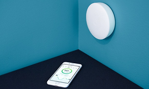 Mobiltelefon som ligger i ett hörn på ett svart golv och blå väggar, med en vit Airthings Wave Plus på ena väggen.