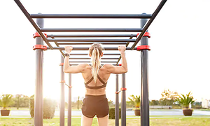Kvinna i svarta shorts och sport bh tränar pull-ups i utegym med sportklocka på.
