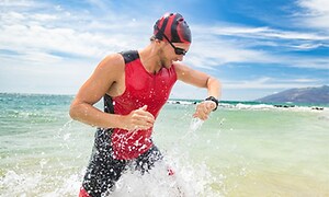 Man i simdräkt, badmössa och simglasögon på väg upp ur vattnet på en strand kollar på sin sportklocka. 